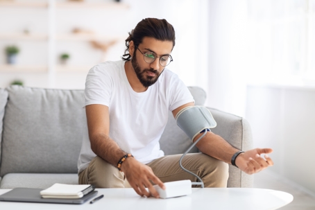 Young arab man sitting on couch, checking blood pressure and taking notes at home.